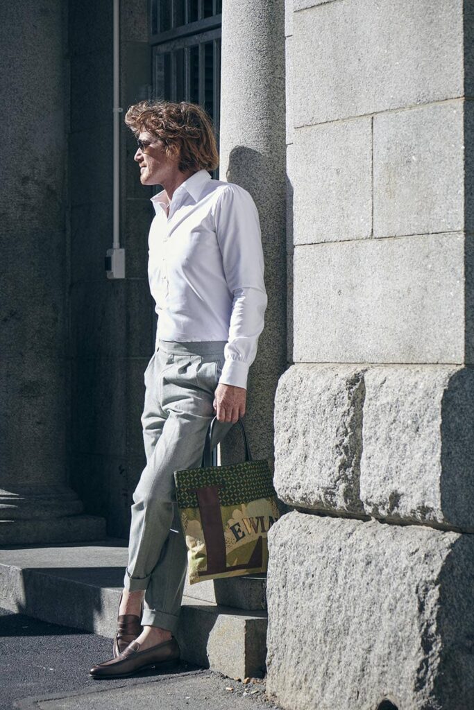 Ein Mann lehnt an einem Haus, im weißen Shirt und Denim Hose